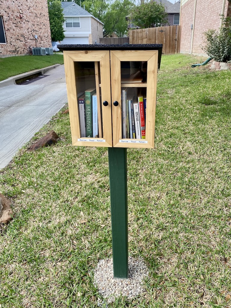 A Little Free Library-style wooden shelf stocked with books, placed outdoors in a neighborhood